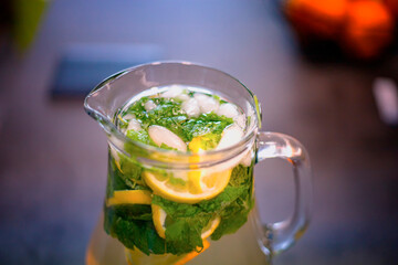 Close up photo of ice cold lemonade with mint and lemon. The pitcher is standing on the kitchen counter. Background is blurred.