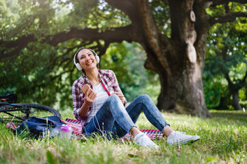 Woman with headphones sitting on a grass in a public park listening music over smartphone singing loud. Positive emotions
