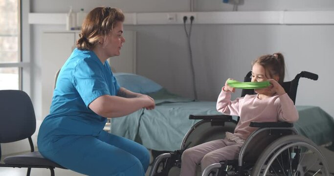 Nurse helping little girl patient in wheelchair during physical exercise with resistance band