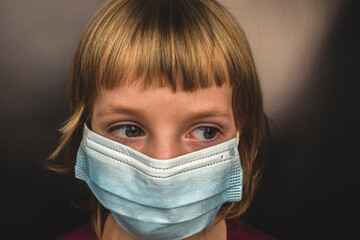 Little blonde girl in a blue mask looks to the side on a black background. Quarantine restrictions and rules for wearing masks for children