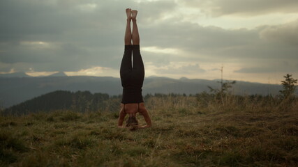 Girl standing on head in mountain landscape. Trainer practicing yoga on ground