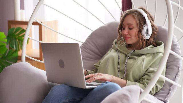 Happy smile woman listen music headphones use laptop typing text message on laptop sitting chair dressed blue jeans oversize hoodie Caucasian female enjoy sound listen podcast audio books video call
