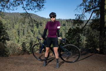 Fototapeta na wymiar Joven mujer ciclista posando con su bicicleta en un mirador con un paisaje boscoso al fondo. Ciclista profesional con casco y gafas de sol.