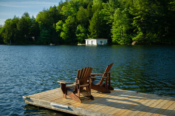Two Adirondack chairs on a wooden dock facing a lake in Muskoka, Ontario Canada during a sunny summer morning. A white boathouse is visible across the water.
