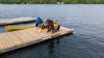 Aerial view of two Adirondack chairs on a wooden dock facing a lake in Muskoka, Ontario Canada. Life jackets and paddles are visible on the chairs. A yellow canoe is tied to the pier.
