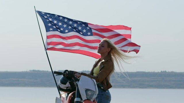 A girl, an FMX motocross racer, wears a helmet on her head, stands next to a motorcycle against the backdrop of a US fdag developing in the wind. Super slow motion, 240 fps.
