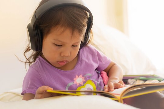 A little girl reading a book at home and listening to music wearing headphones. Audio books.