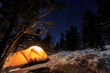 Overnight stay at the top of the winter mountain. An orange tent, illuminated from the inside, stands at the edge of the forest in the middle of the night in the snow.