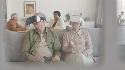 Medium long of grey-haired man and woman wearing virtual reality headsets, sitting on couch in living room, moving heads sideways, up and down, talking, smiling - Powered by Adobe