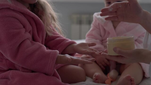 Authentic cute young woman applying face mask of baby girl. mom with 2 children daughters different ages in bathrobes after bath on white bed oposite window. Spa, motherhood, childhood, family concept