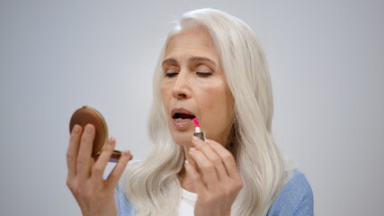 Senior woman putting on makeup in studio. Mature lady looking to mirror indoor.