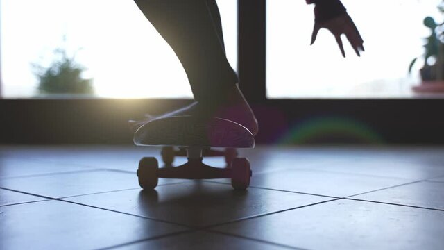 A backlit shot of a barefoot young man or woman stays in balance on a skateboard from a standstill in front of the window at home at sunset. 