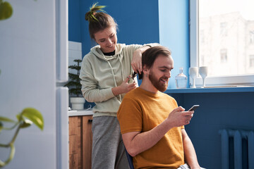 Loving couple spending time at the kitchen while woman cutting hair of her husband