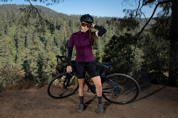 Joven mujer ciclista  posando con su bicicleta en un mirador con un paisaje  boscoso al fondo. ...