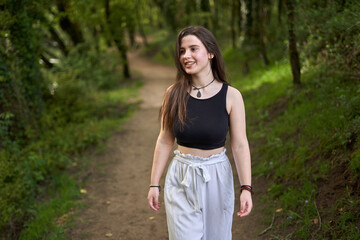 Fototapeta premium Young girl with dark hair and light skin in a black top, slouchy white pants, black and white sneakers and necklace `walking along a dirt path in lush green forest. woman in nature. teenager hiking