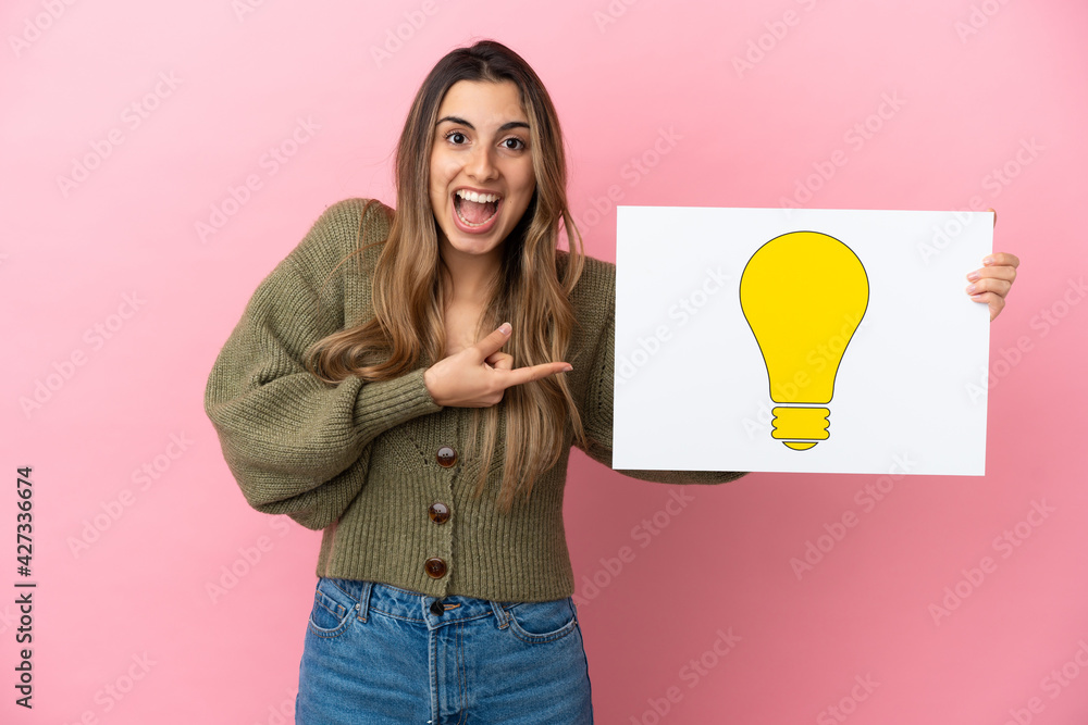 Wall mural Young caucasian woman isolated on pink background holding a placard with bulb icon and pointing it - Wall murals