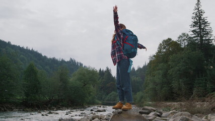 Happy girl standing on rock at river bank. Female hiker raising hands in air