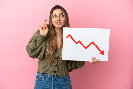 Young caucasian woman isolated on pink background holding a sign with a decreasing statistics arrow symbol with fingers crossing