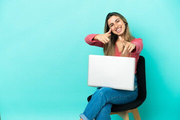 Young caucasian woman sitting on a chair with her pc isolated on blue background pointing to the front and smiling