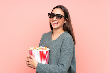Young caucasian woman isolated on pink background with 3d glasses and holding a big bucket of popcorns