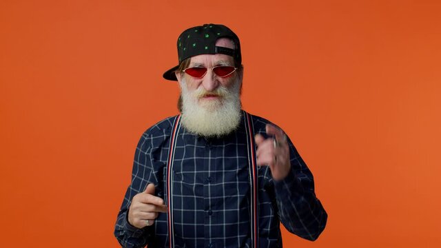 Aggression. Angry elderly cool bearded gray-haired man in cap shakes hands with rage, furious complains about something has quarrel with someone. Senior old grandfather isolated on orange background.