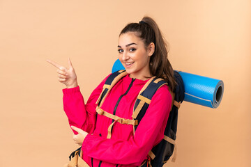 Young mountaineer girl with a big backpacker isolated on beige background pointing finger to the side