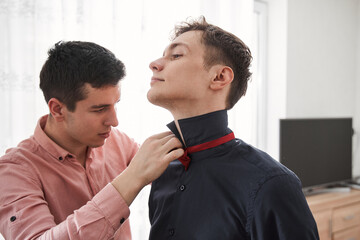 Gay standing and helping to put on a bowtie to his boyfriend while at home