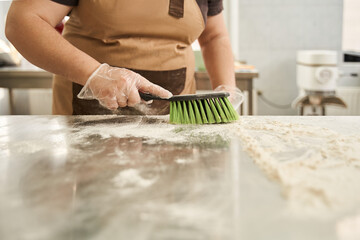 Female baker wearing protective gloves cleaning with brush table of the flour