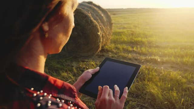 Smart technologies in agriculture. A pretty young woman farmer uses a tablet near a haystack in a mown field at sunset, planning and running an agribusiness using smart technologies.