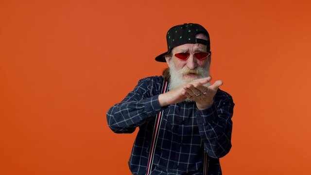Mature old bearded grandfather in sunglasses and cap showing wasting or throwing money around hand gesture, more tips dreaming about big profit body language. Senior man isolated on orange background
