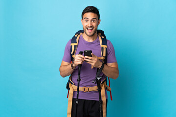 Young caucasian man with backpack and trekking poles isolated on blue background surprised and sending a message