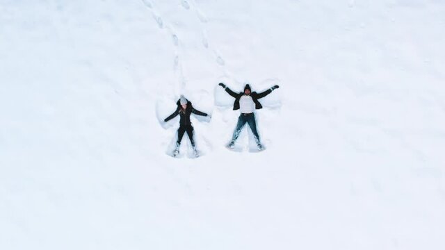 Slow-motion drone video of a happy young couple having fun during winter laying down on snow moving arms and legs. High angle view