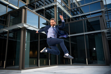 Fototapeta premium Funny positive businessman or worker in suit jumping in front of an office glass building. Excited business people.