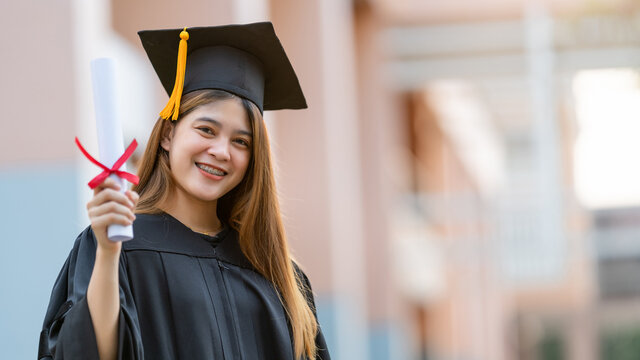 A young happy Asian woman university graduate in graduation gown and mortarboard holds a degree certificate celebrates education achievement in the university campus.  Education stock photo