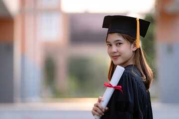 A young happy Asian woman university graduate in graduation gown and mortarboard holds a degree...