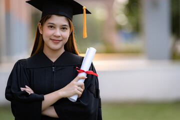 A young happy Asian woman university graduate in graduation gown and mortarboard holds a degree...