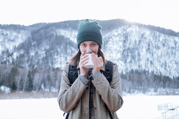 Fototapeta na wymiar A young tourist woman drinking a drink from a white cup on the mountain. Hiker woman enjoying winter mountains. Outdoor activities in the mountains. Traveler with backpack