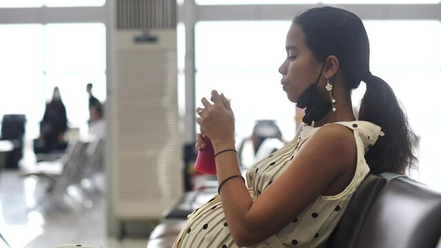 Pregnant woman in mask at aiport departure area drinking a healthy juice.