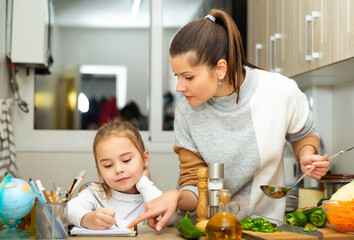 Little girl writing school lesson during mother cooking at kitchen