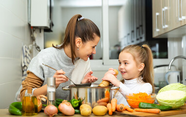 Positive mother and little daughter tasting soup together at home