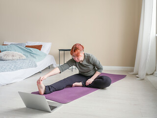 Young woman doing stretching looking into laptop. Bedroom home interior. Concept of online yoga training and healthy life in self-isolation