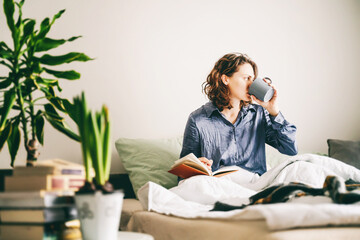 Young cheerful woman sitting on bed writing in diary drinking morning coffee