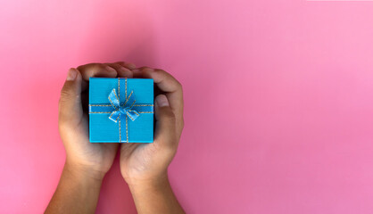 Top view image kid child's hands holding present blue color small gift box wrapped with blue ribbon  for birthday, Valentine, Mother day or wedding put on pink pastel table top view background.