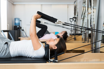 Active Hispanic girl enjoying pilates classes in modern fitness studio with group, doing exercises on reformer