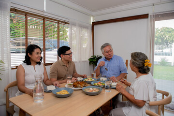 Asian family having dinner together at home. Smiling adult couple with senior parents enjoy eating and sharing thai food on dining table. Happy family spending time together on weekend vacation.