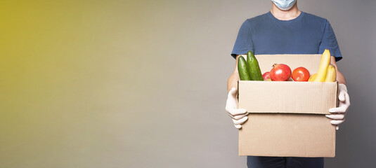 Male courier in mask.Man with cardboard box of groceries, fast and safe food delivery.Deliveryman with products on yellow and grey background banner.