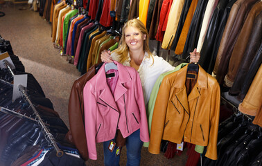 Attractive young woman choosing leather jacket on racks in clothes store