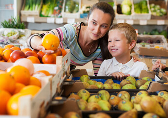 Cheerful brunette mother with little boy buying oranges at store