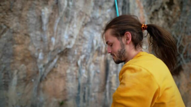 slow motion of joyful handsome man rock climber in yellow sweatshirt with long hair hanging on rope and give high five after successful ascent of route on cliff. 4k footage