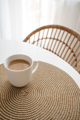 White cup of coffee on wicker place mat. Background with white table and rattan chair. Boho style...